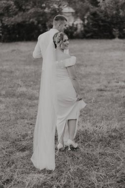 Gelin ve damat tarlada duruyorlar, gelin peçe takıyor ve damat takım elbise giyiyor.