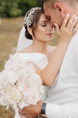 Bir gelin ve damat bir tarlada birbirlerini kucaklıyor. Gelin beyaz bir elbise ve çiçekli bir saç bandı takıyor.