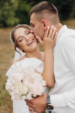 Bir gelin ve damat birbirlerinin dudaklarını öpüyor. Gelin elinde bir buket çiçek tutuyor.