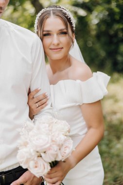 Bir gelin ve damat parkta fotoğraf çektirmek için poz veriyorlar. Gelin bir buket çiçek tutuyor ve damat da onu tutuyor. Sahne mutlu ve romantik.