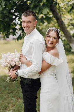 Bir gelin ve damat bir buket çiçekli fotoğraf için poz veriyorlar. Gelin beyaz bir elbise ve duvak takarken damat beyaz gömlek ve siyah pantolon giyiyor. İkisi de gülümsüyor.