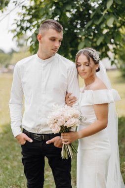 Bir gelin ve damat tarlada fotoğraf çektirmek için poz veriyorlar. Gelin bir buket çiçek tutuyor ve damat elini tutuyor. Sahne mutlu ve romantik.