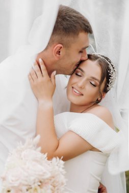 Bir gelin ve damat birbirlerinin alnını öpüyor. Gelin beyaz bir elbise giyiyor ve damat beyaz bir gömlek giyiyor.