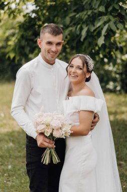 Bir gelin ve damat bir buket çiçekli fotoğraf için poz veriyorlar. Gelin beyaz bir elbise ve duvak takarken damat beyaz bir gömlek giyiyor. Sahne mutlu ve neşeli.