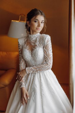 Uzun beyaz elbiseli bir kadın resim için poz veriyor. Kolye ve yüzük takıyor. Elbise uzun ve bir sürü parıltısı var.