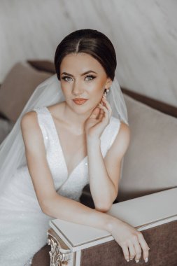 Beyaz elbiseli bir kadın, elleri kalçasında bir kanepede oturuyor. Saçında bir peçe var ve bir kolye takıyor. Zarafet ve incelik kavramı