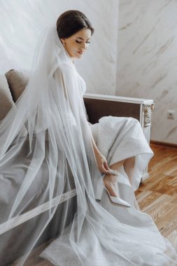 Bir gelin kafasında peçeyle bir kanepede oturuyor. Beyaz bir elbise ve beyaz ayakkabılar giyiyor.