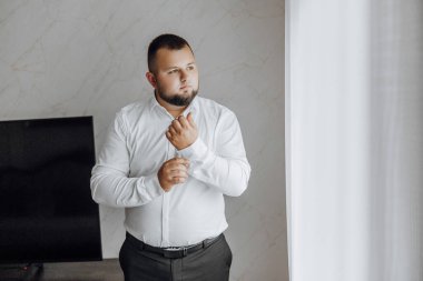 Beyaz gömlekli bir adam pencerenin önünde giyiniyor. Siyah kuşak ve siyah gömlek giyiyor.