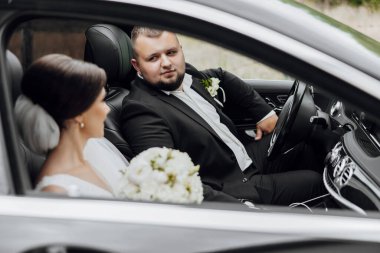 Bir adam ve bir kadın arabada oturuyorlar. Adam takım elbise ve kravat takıyor. Adamın elinde bir buket çiçek var.