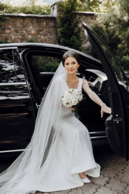 Gelinlikli bir kadın siyah bir arabanın arkasında oturuyor. Elinde bir buket çiçek ve duvak tutuyor. Sahne muhtemelen bir düğün ya da özel bir olay.