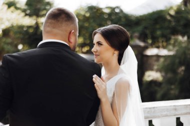 Gelin ve damat balkonda duruyorlar, gelin beyaz bir elbise giyiyor ve damat siyah bir takım elbise giyiyor.