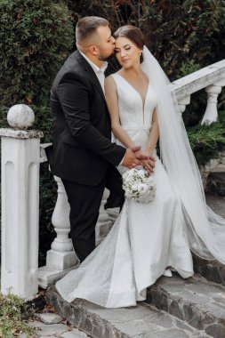 Bir gelin ve damat merdivenlerde fotoğraf çektirmek için poz veriyorlar. Gelin beyaz bir elbise giyiyor ve damat siyah bir takım elbise giyiyor. Sahne mutlu ve romantik.