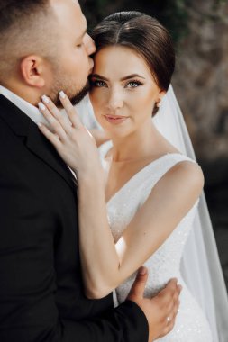 Gelin ve damat birbirine sarılıyor, gelin beyaz bir elbise giyiyor. Adam kadının dudaklarını öpüyor.