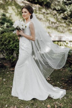 Bir gelin, elinde bir buket beyaz çiçek ve peçeyle bir tarlada duruyor.