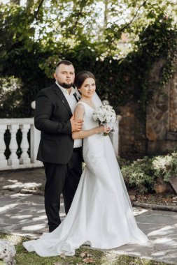 Bir gelin ve damat taş bir duvarın önünde poz veriyorlar. Gelin elinde bir buket çiçek tutuyor.