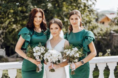 Yeşil elbiseli üç kadın ellerinde çiçek buketleri tutuyor. Gülümsüyorlar ve poz veriyorlar. Sahne neşeli ve kutlama dolu.