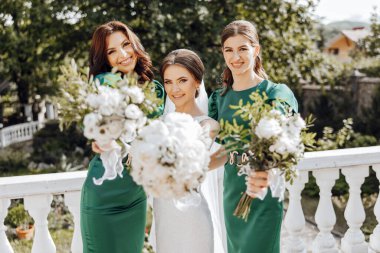 Yeşil elbiseli üç kadın ellerinde beyaz çiçekler tutuyor. Gülümsüyorlar ve resim için poz veriyorlar. Sahne mutlu ve kutlama dolu, çünkü kadınlar büyük ihtimalle bir düğüne ya da diğer özel etkinliklere katılıyorlar.