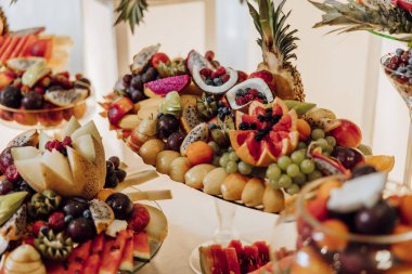 Elma, portakal ve üzüm gibi çeşitli meyvelerle dolu bir meyve tabağı. Tabak bir masaya konur ve diğer meyve tabakları ile çevrilidir.