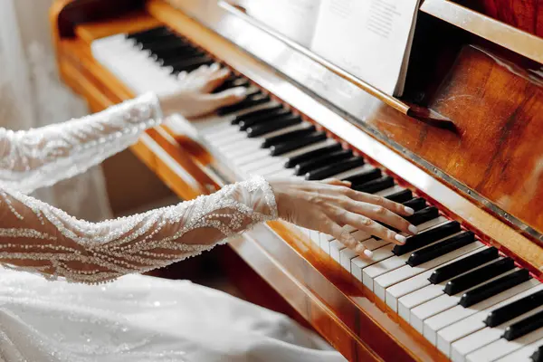 A woman is playing the piano with her hands on the keys. The piano is an old model and the woman is wearing a white dress. Scene is elegant and sophisticated