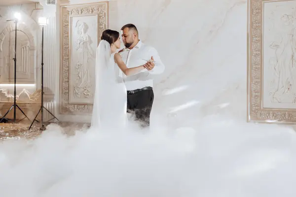 A bride and groom are dancing in a misty room. The bride is wearing a white dress and the groom is wearing a white shirt