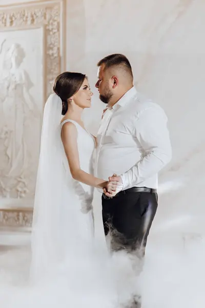 A bride and groom are dancing in a misty room. The bride is wearing a white dress and the groom is wearing a white shirt and black pants