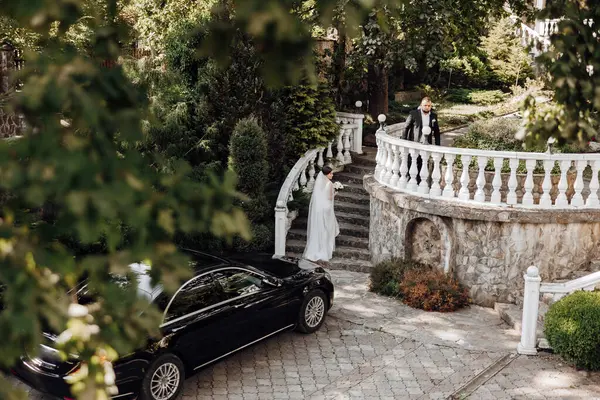 黒い車の前に白い階段を歩いている花嫁と新郎 シーンは庭に設定され 白い柵といくつかの鉢植え植物があります シーンはロマンチックで親密です ロイヤリティフリーのストック写真