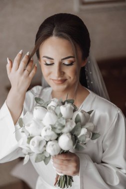 Bir kadın elinde bir buket beyaz çiçek tutuyor. Beyaz bir bornoz giyiyor ve saçını arkaya çekiyor. Çiçekler bir vazoya konur ve onun önüne yerleştirilir.
