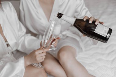 İki kadın bir yatakta oturuyor, biri bardağa şampanya dolduruyor. Sahne kutlama ve romantiktir.