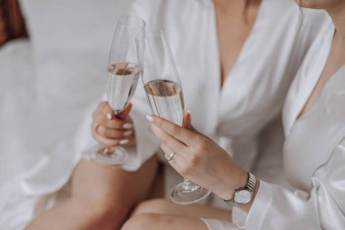 İki kadın bir yatakta oturuyor, ellerinde şampanya bardakları ve gülümsüyorlar. Sahne kutlama ve mutluluk dolu.