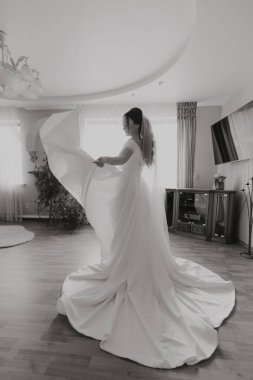 Beyaz elbiseli bir kadın büyük aynalı bir odada duruyor. O, beyaz bir elbise tutuyor ve bir düğün için hazırlanıyor. Oda saksı bitkisi ve vazoyla süslenmiş.