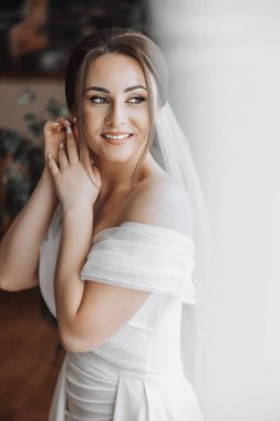 Beyaz bir elbise ve duvak takan bir kadın var. Gülümsüyor ve kameraya bakıyor.