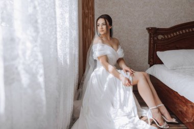 Beyaz elbiseli bir kadın yatakta oturuyor. Peçe takıyor ve bacak bacak üstüne atıyor.