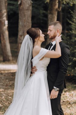 Gelin ve damat ormanda duruyorlar, gelin beyaz bir elbise ve damat siyah bir takım elbise giyiyor.