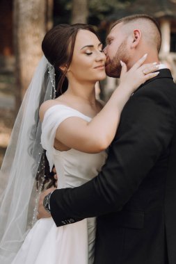 Bir gelin ve damat birbirlerinin dudaklarını öpüyor. Gelin beyaz bir elbise giyiyor ve damat siyah bir takım giyiyor.