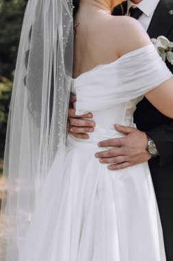 Bir gelin ve damat düğün fotoğrafında birbirlerine sarılıyorlar. Gelin peçeli beyaz bir elbise giyiyor ve damat takım elbise giyiyor. Fotoğraf bir aşk anını yakalar.