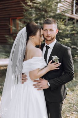 Bir gelin ve damat bir ağacın önünde fotoğraf için poz veriyorlar. Gelin beyaz bir elbise giyiyor ve damat siyah bir takım elbise giyiyor. İkisi de gülümsüyor ve mutlu görünüyorlar.