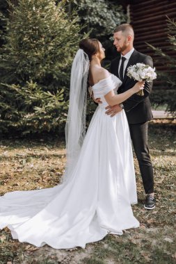 Gelin ve damat tarlada durur, gelin beyaz bir elbise giyer ve damat siyah bir takım giyer.