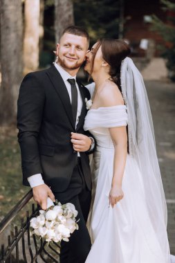 Bir gelin ve damat birbirlerinin yanaklarını öpüyor. Gelin beyaz bir elbise giyiyor ve damat siyah bir takım giyiyor.