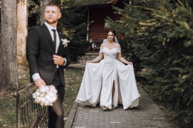 Gelin ve damat, gelin elinde buketle bir patikada yürüyorlar. Sahne romantik ve mutlu, çift düğünlerine hazırlanıyor.