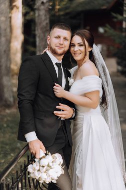 Bir gelin ve damat çitin önünde fotoğraf için poz veriyorlar. Gelin beyaz bir elbise giyiyor ve damat siyah bir takım elbise giyiyor. Bir buket çiçek ve çiçek tutuyorlar.