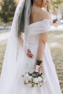 Beyaz elbiseli bir kadın elinde bir buket çiçek tutuyor. Duvak takıyor ve kollarını kavuşturuyor. Sahne bir parkta geçiyor, arka planda bir bank var.