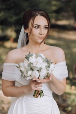 Bir kadın elinde bir buket çiçek tutuyor. Beyaz bir elbise giyiyor ve saçları topuz şeklinde.
