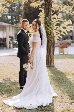 Bir gelin ve damat parkta fotoğraf için poz veriyorlar. Gelin beyaz bir elbise giyiyor ve damat takım elbise giyerken elinde bir buket tutuyor. Sahne aydınlık ve neşeli.