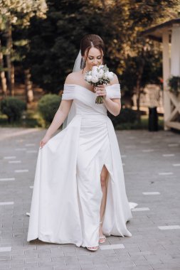 Bir gelin elinde bir buket çiçekle kaldırımda yürüyor. Sahne parlak ve neşeli, gelin mutlu ve kendinden emin görünüyor.