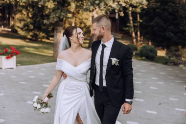 Bir gelin ve damat yolda yürüyor, hem gülümsüyor hem de ellerinde çiçekler var. Sahne mutlu ve kutlama dolu, çünkü çift yeni evli.