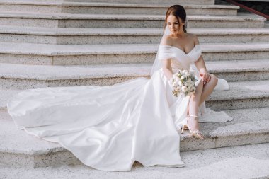 Beyaz gelinlikli bir kadın elinde bir buket çiçekle merdivenlerde oturuyor.