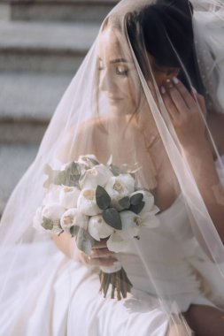 Bir gelin basamakta oturuyor ve yüzünü örten bir peçeyle. Kucağında bir buket çiçek tutuyor.