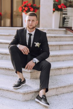Takım elbiseli ve kravatlı bir adam merdivenlerde oturur. Bir saat ve yüzük takıyor.