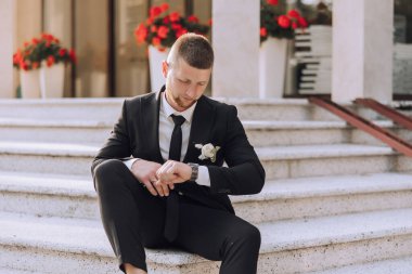 Takım elbiseli ve kravatlı bir adam merdivenlerde oturuyor. Bir saat takıyor ve saati kontrol ediyor. Adam takım elbise giydiği için resmiyet ve profesyonellik kavramı.