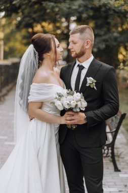 Bir gelin ve damat bir bankın önünde ellerinde bir buket ve vazoyla duruyorlar.